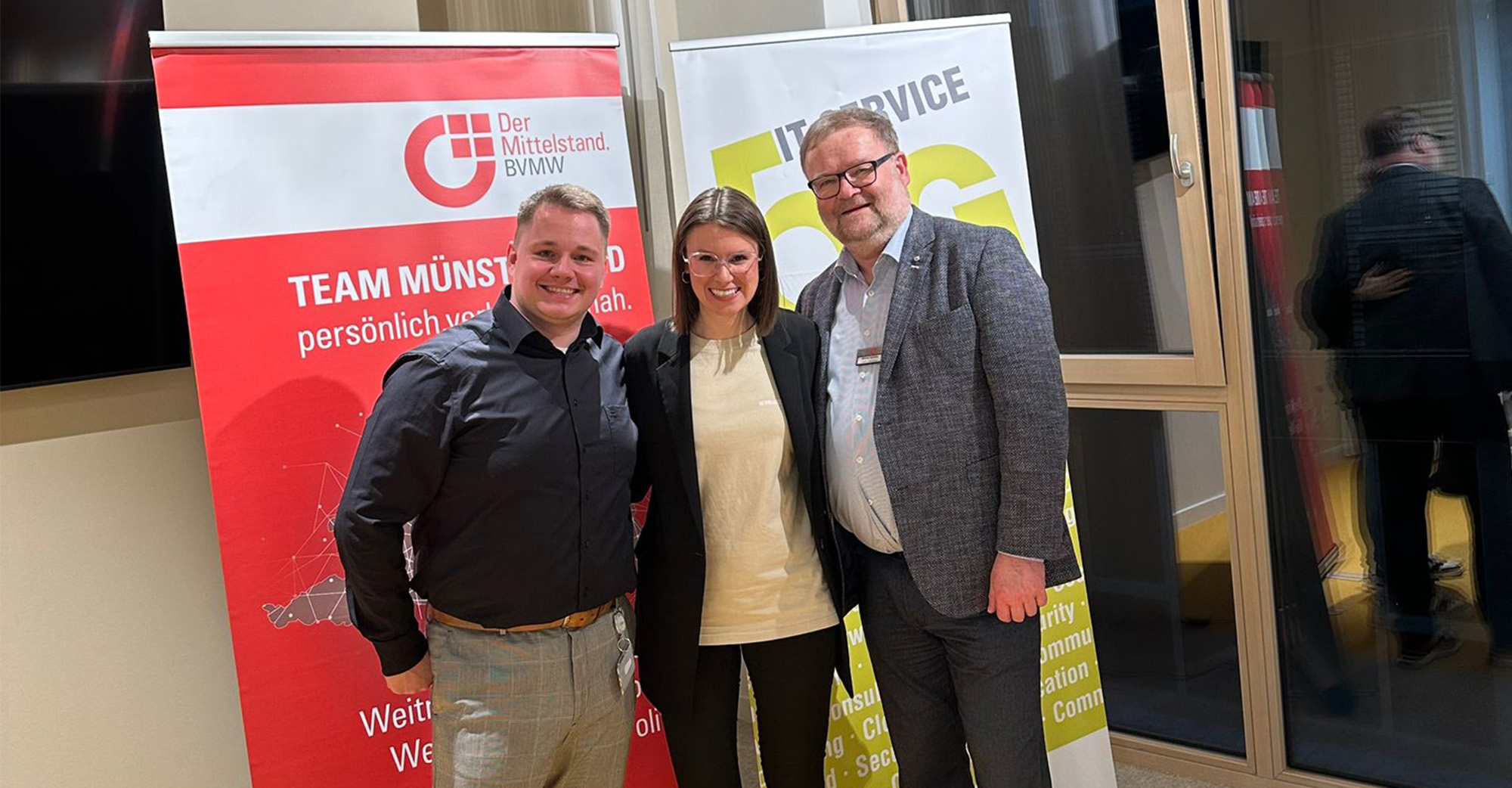Drei Personen stehen eng zusammen und lächeln in die Kamera bei einer Indoor Netzwerkveranstaltung. Hinter ihnen stehen zwei Roll up Banner, links ein rotes Banner mit dem Logo und Text „Der Mittelstand BVMW“ und „TEAM MÜNSTERLAND“, rechts ein weißes Banner mit großem Schriftzug „IT SERVICE“.