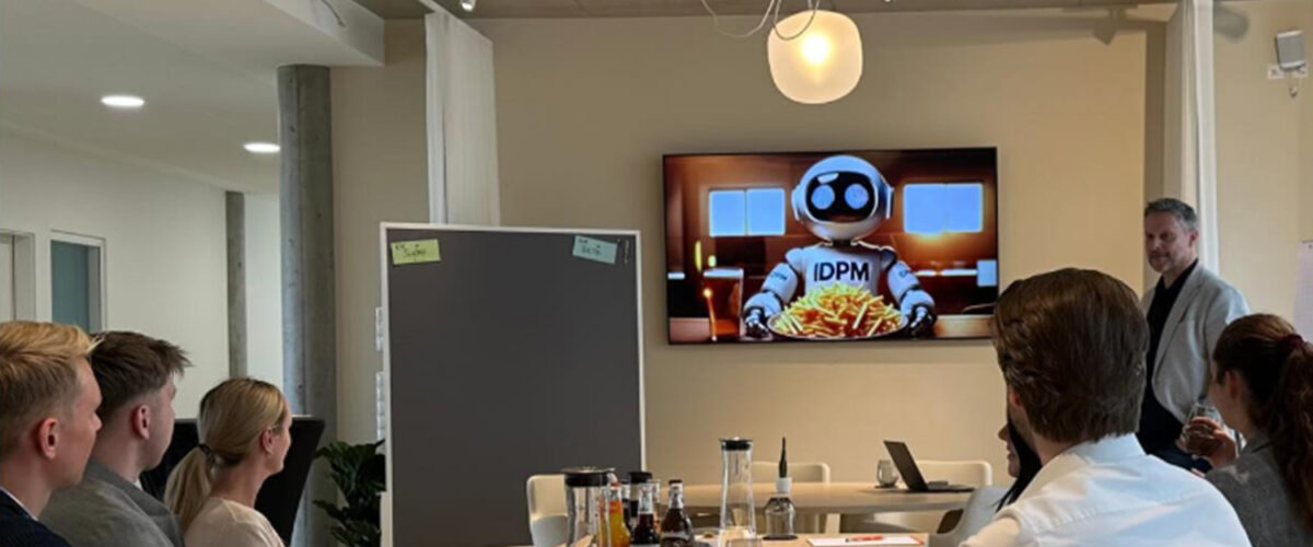 Teilnehmer sitzen an einem Tisch im modernen IDPM Marktplatz und verfolgen einen Vortrag und schauen interessiert auf einen TV. Auf dem Bildschirm ist ein Roboter mit Pommes und dem Schriftzug IDPM zu sehen.