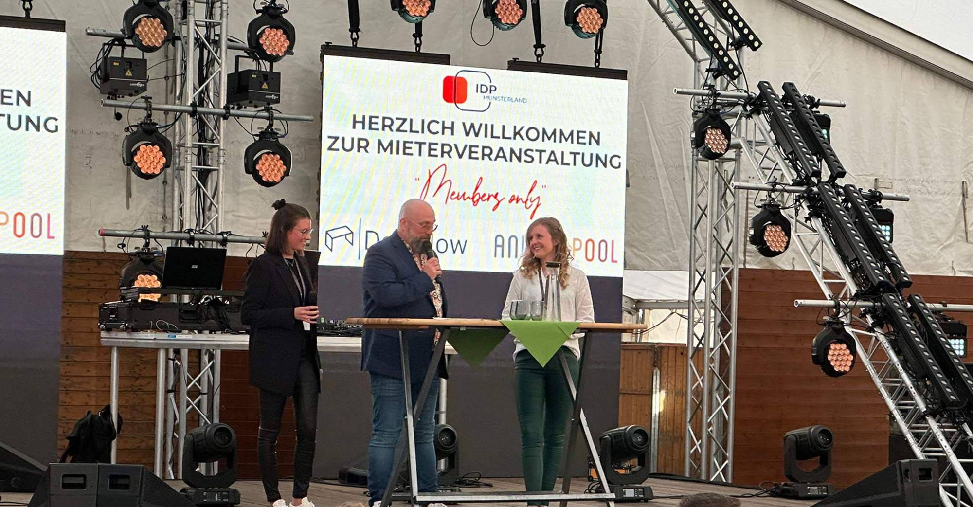 Bühne in einem großen Zelt bei einer Mieterveranstaltung mit Lichttraversen, Scheinwerfern und zwei großen Bildschirmen im Hintergrund. Auf dem mittleren Screen steht „Herzlich willkommen zur Mieterveranstaltung“ mit IDP Münsterland, davor stehen drei Personen an einem hohen Stehtisch, eine Person spricht ins Mikrofon, zwei weitere hören zu. Im Vordergrund sitzt Publikum an Tischen und schaut zur Bühne.