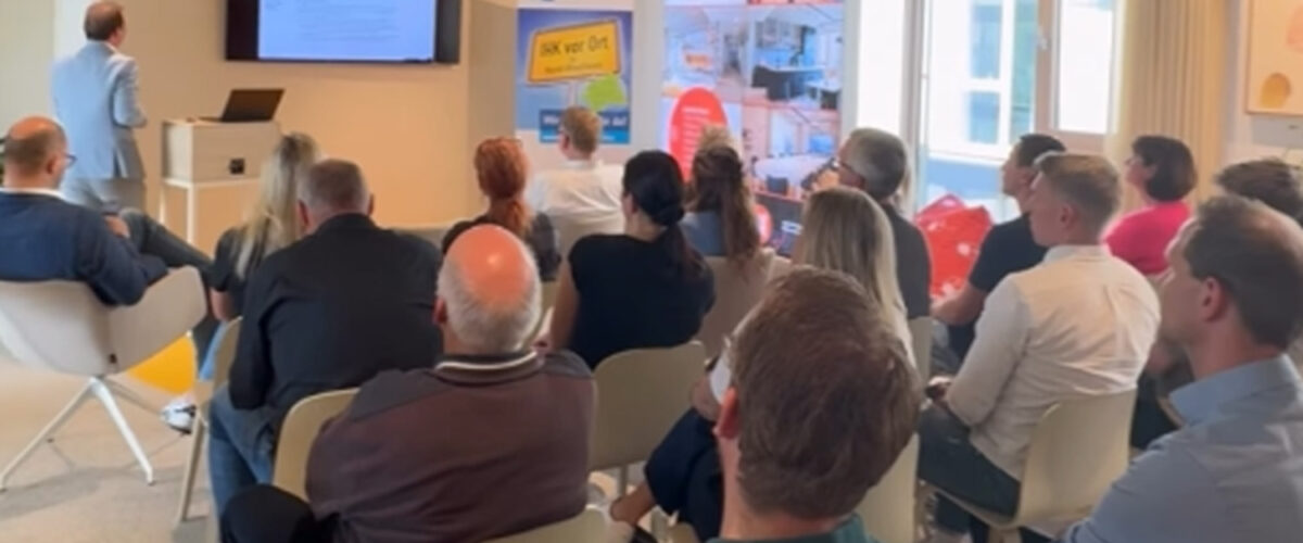 Publikum sitzt im Marktplatz vom IDPM und verfolgt einen Vortrag, der Referent präsentiert Folien auf einem großen Bildschirm.