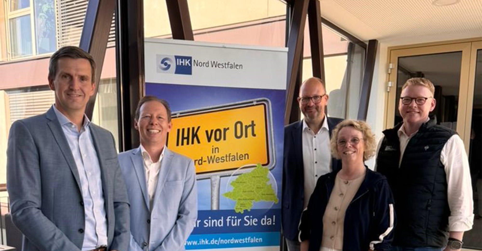Fünf Personen stehen in einem hellen Innenbereich mit großen Fensterflächen und dunklen Stahlträgern und posieren für ein Foto. In der Mitte steht ein Roll up Banner mit der Aufschrift „IHK Nord Westfalen“ und „IHK vor Ort in Nord Westfalen“, darunter der Satz „Wir sind für Sie da“.