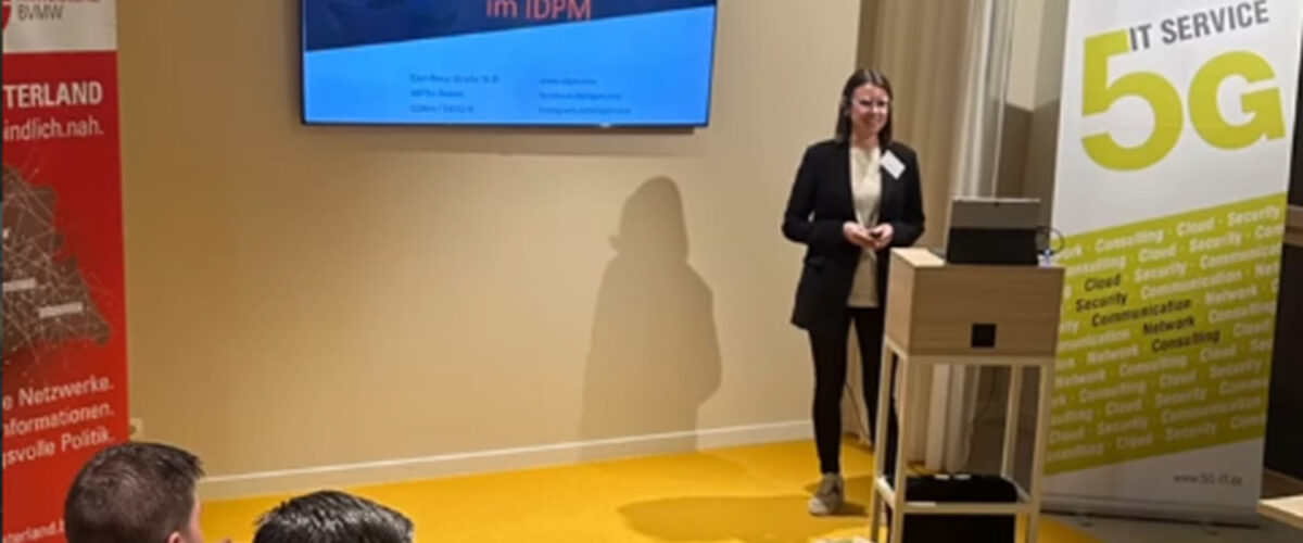 Referentin steht bei einer Veranstaltung im IDPM vor einer Begrüßungsfolie mit dem Text Herzlich Willkommen im IDPM, im Vordergrund sitzen Teilnehmende und hören interessiert zu.